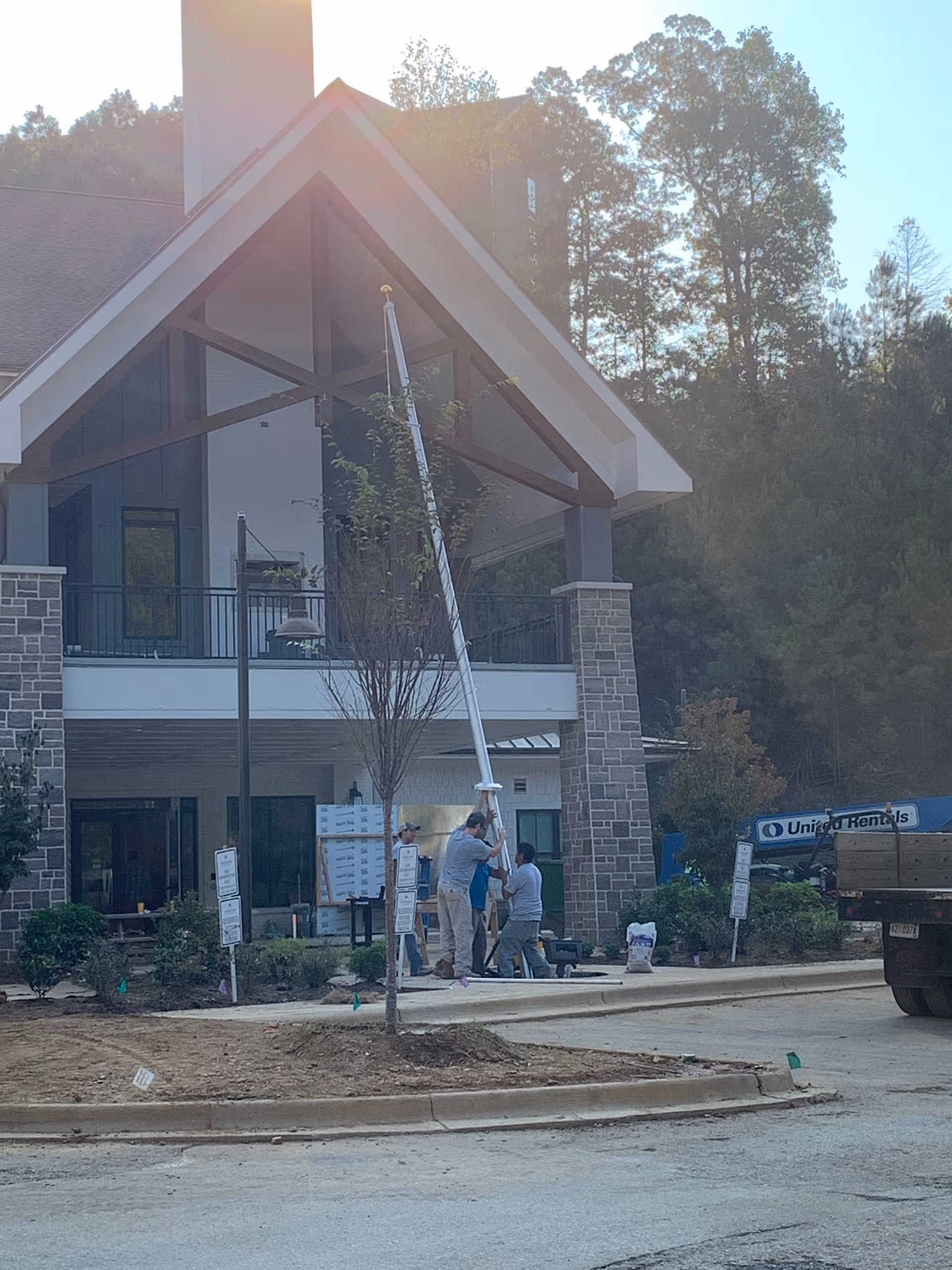 Workers installing a tall pole at the covered stone entrance of a multi-story building with trees in the background.