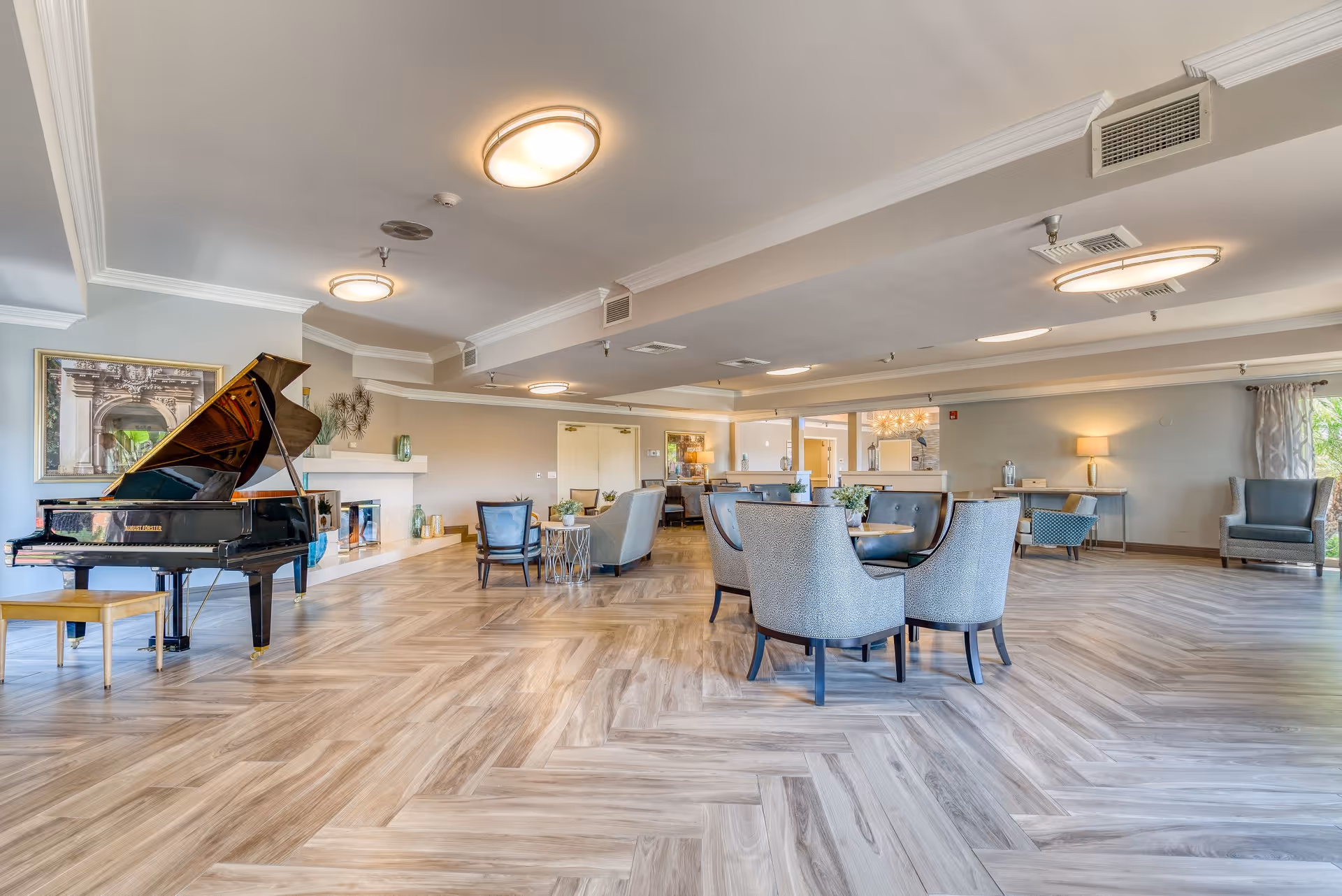 Bright, spacious senior living common room featuring a grand piano, multiple seating areas, and patterned wood floors.
