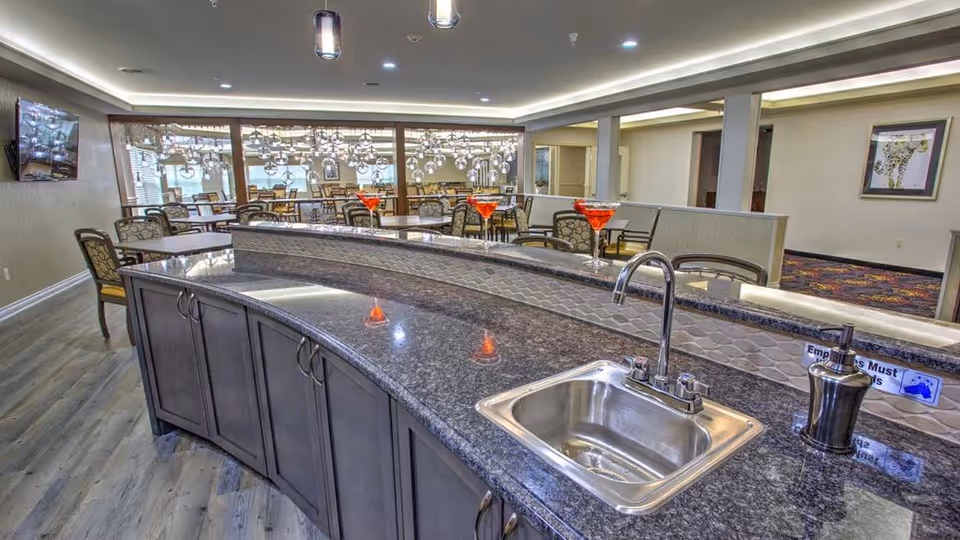 Interior view of a dining area in a senior living facility featuring a curved granite countertop with a built-in sink and soap dispenser. Behind the counter are multiple tables and chairs arranged for dining, with decorative hanging lights and a carpeted area visible in the background.