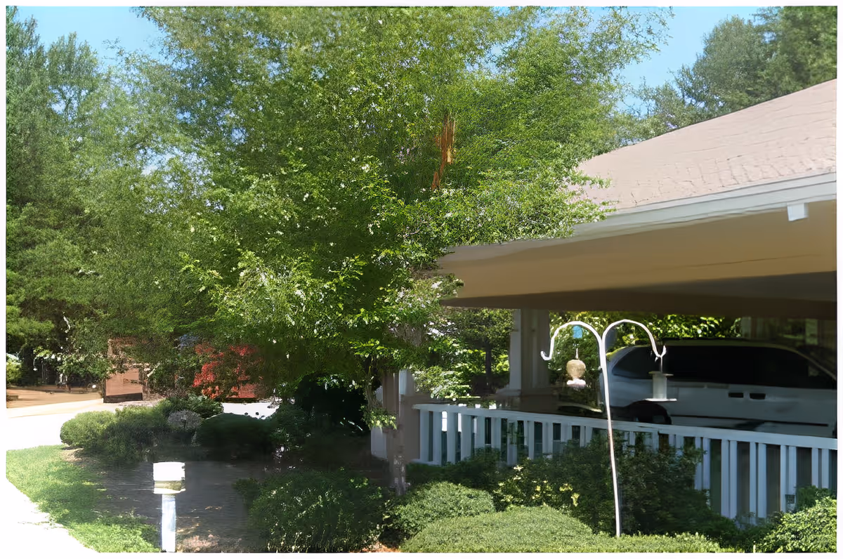 Outdoor view of a senior living facility showing a covered parking area with a white vehicle parked underneath. Surrounding the parking area are green bushes and trees with dense foliage. A bird feeder is hanging on a white metal stand near the bushes.