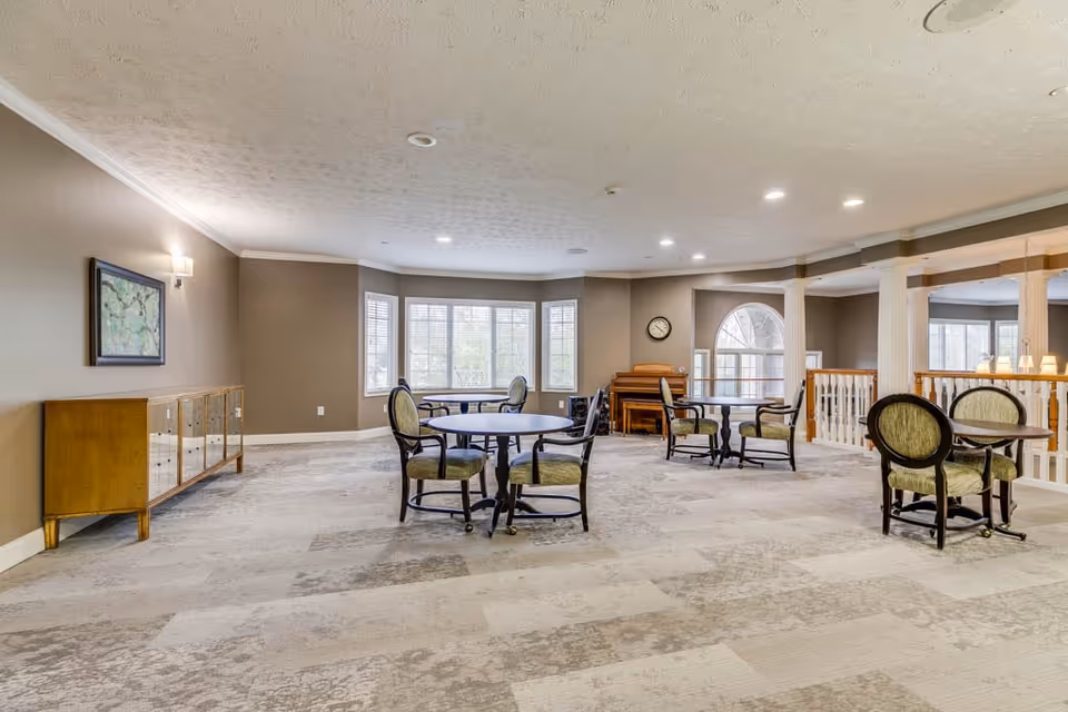 A spacious, well-lit common area with several round tables and chairs arranged for seating. The room features large windows allowing natural light, a piano against the far wall, a clock above the piano, and a sideboard with a framed picture on the left wall. The area has beige walls, patterned carpet flooring, and white ceiling with recessed lighting.