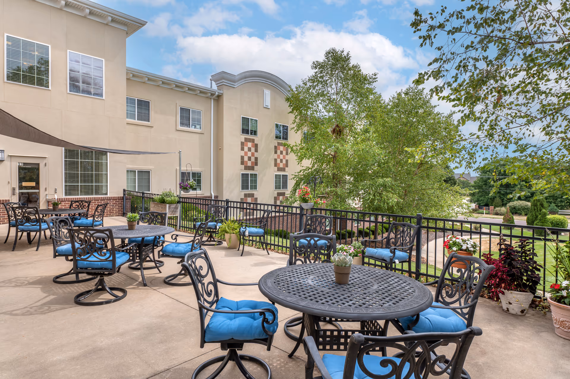 Outdoor patio area at Brookdale Overland Park with black metal tables and chairs featuring blue cushions. The patio is surrounded by a black metal railing and decorated with potted plants and flowers. The building exterior is beige with multiple windows, and there are trees and greenery in the background under a partly cloudy sky.