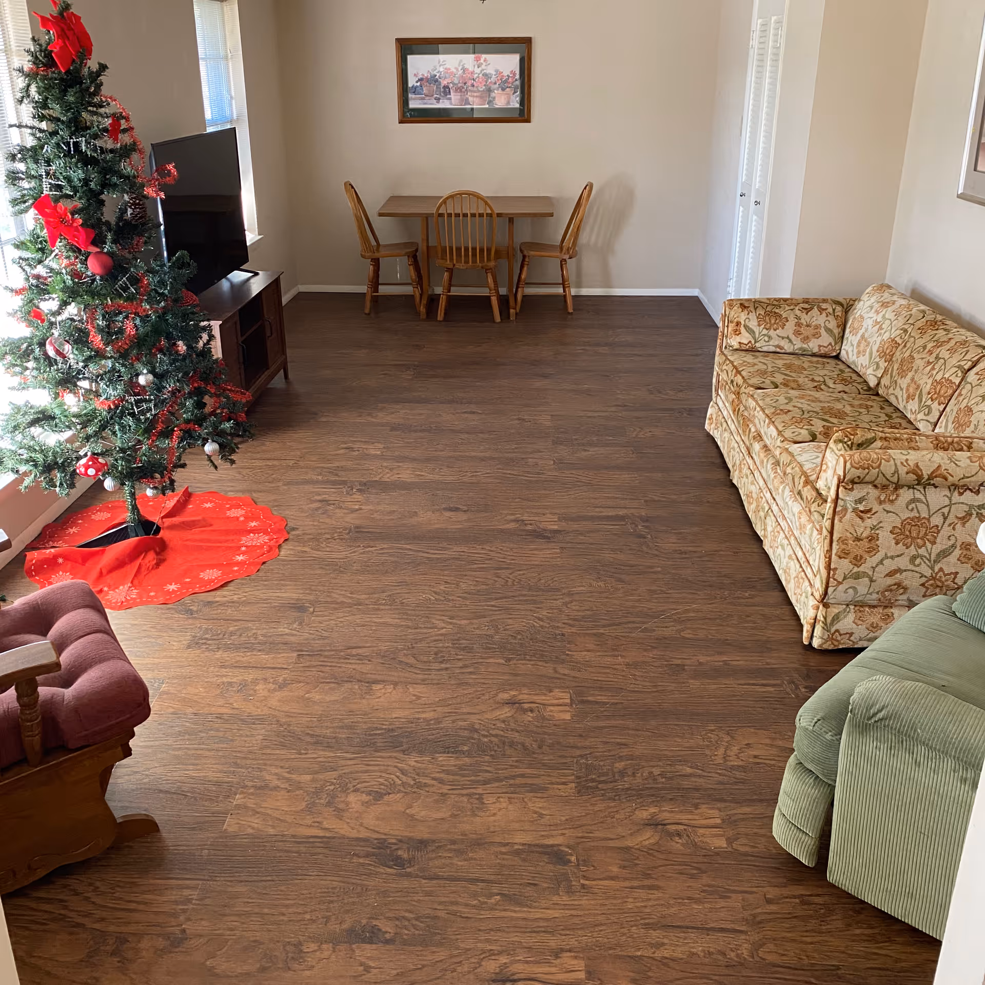 A living room with wood flooring featuring a decorated Christmas tree, a TV stand, a small dining table with chairs, and a floral couch.