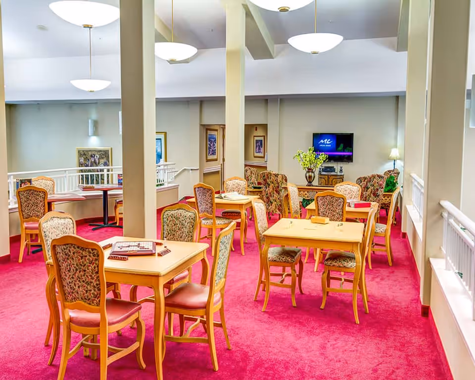 Bright communal dining/activity room with multiple wooden tables and upholstered chairs on pink carpet and a TV seating area in the back.