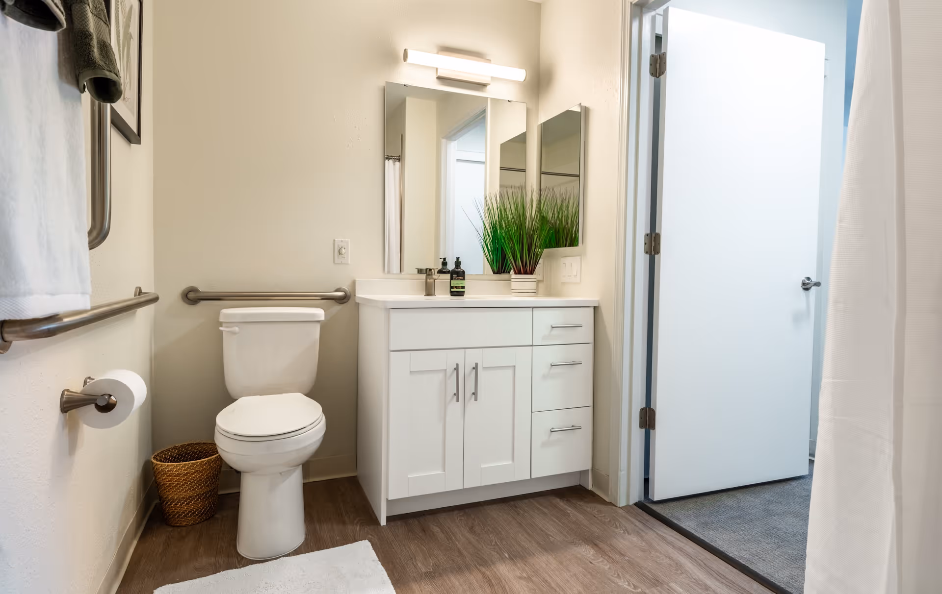 A clean and modern bathroom in Curtis Creek Senior Living featuring a white toilet with grab bars on the walls, a white vanity with a sink and mirror above it, a small potted plant on the vanity, a towel hanging on a rail, a waste basket, and a white door leading to another room.