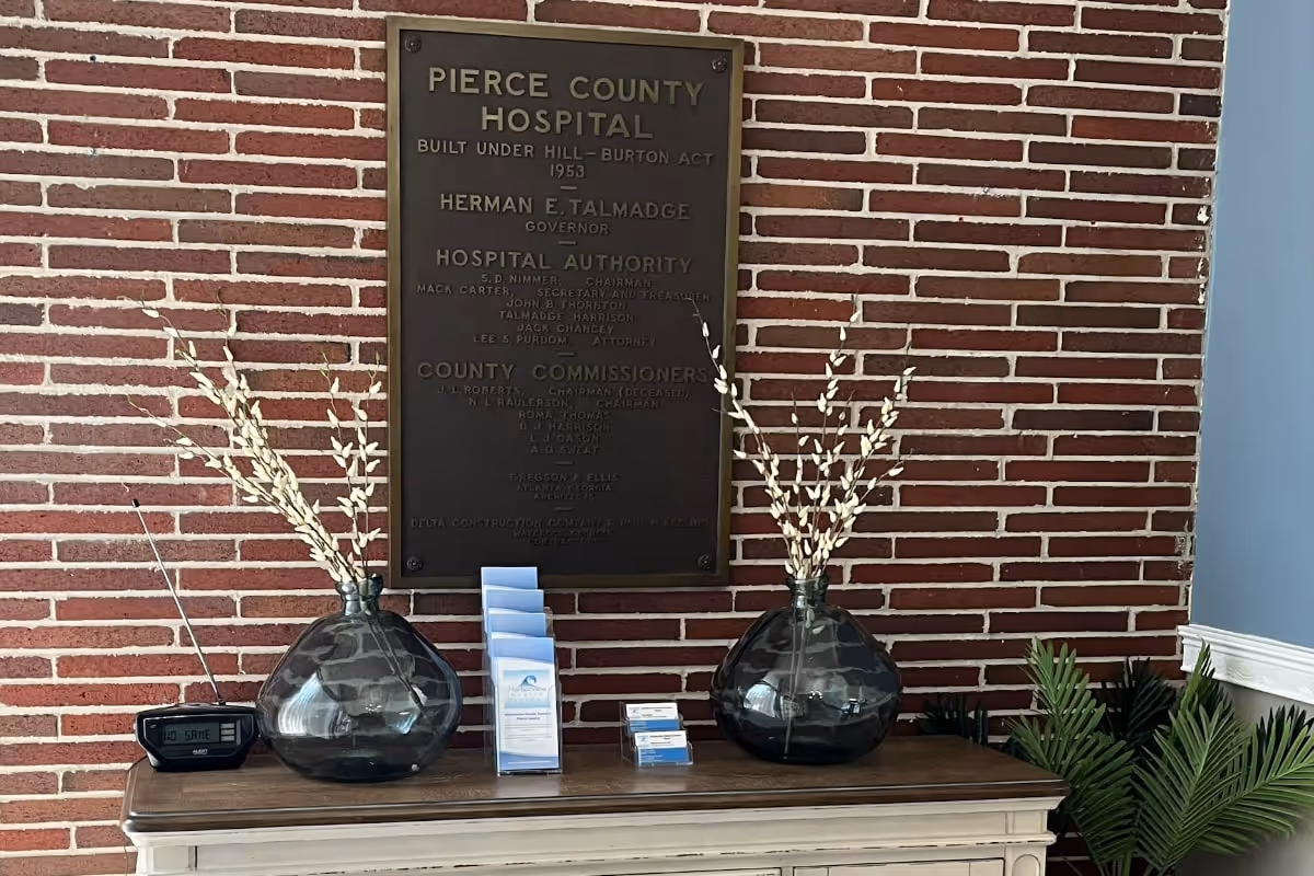 A lobby table against a red brick wall displays a bronze 'Pierce County Hospital' plaque above two glass vases with branches and an information brochure stand.
