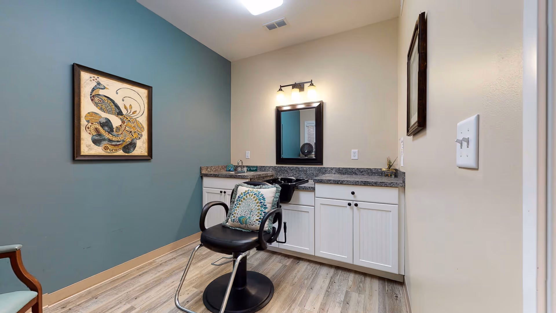 Salon-style room with a black styling chair in front of a countertop sink and mirror against a teal accent wall with framed artwork.