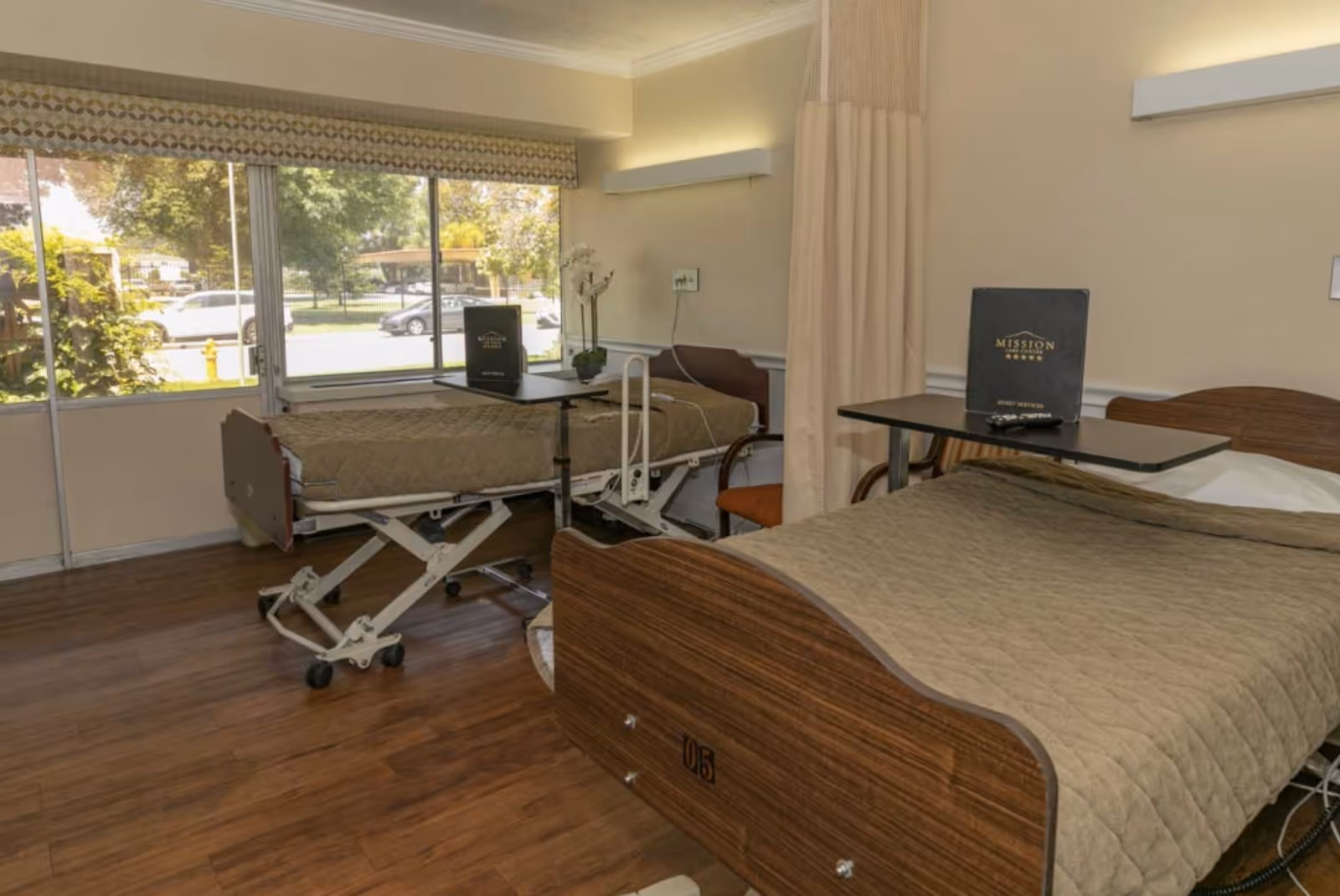 A room with two hospital-style beds, each with a beige quilted blanket and an overbed table. There is a large window with a view of trees and parked cars outside. The room has wooden flooring and a privacy curtain between the beds.