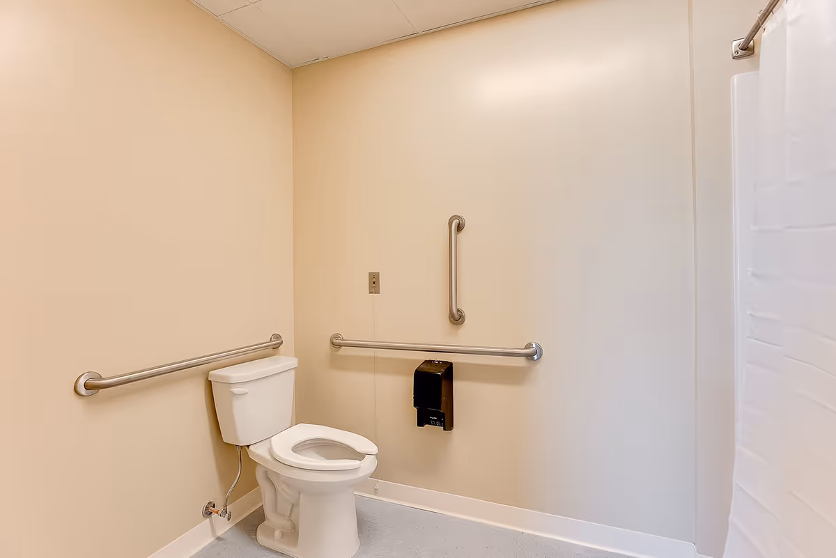 A clean, accessible bathroom with beige walls, a white toilet equipped with safety grab bars on the side and back walls, and a white shower curtain partially visible on the right side.
