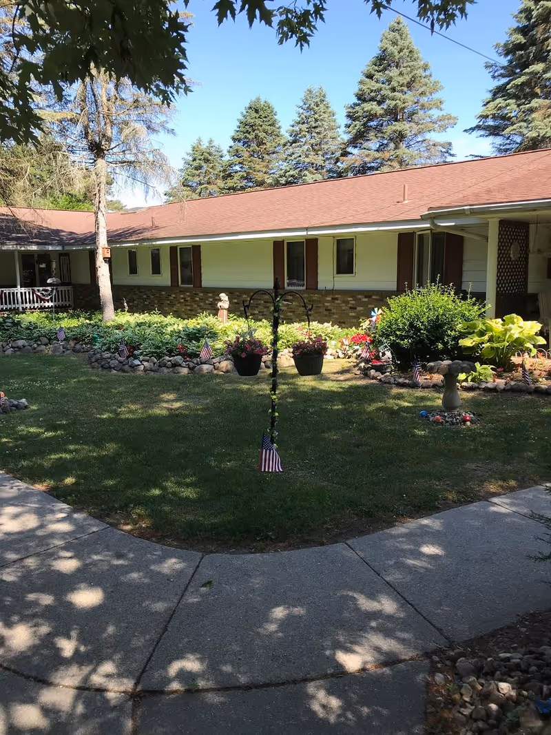 A sunny outdoor courtyard area at Pine Haven Senior Assisted Living with a well-maintained lawn, flower beds bordered by rocks, hanging flower pots, small American flags, and a birdbath. The building with a red roof and white siding is visible in the background, surrounded by tall evergreen trees.