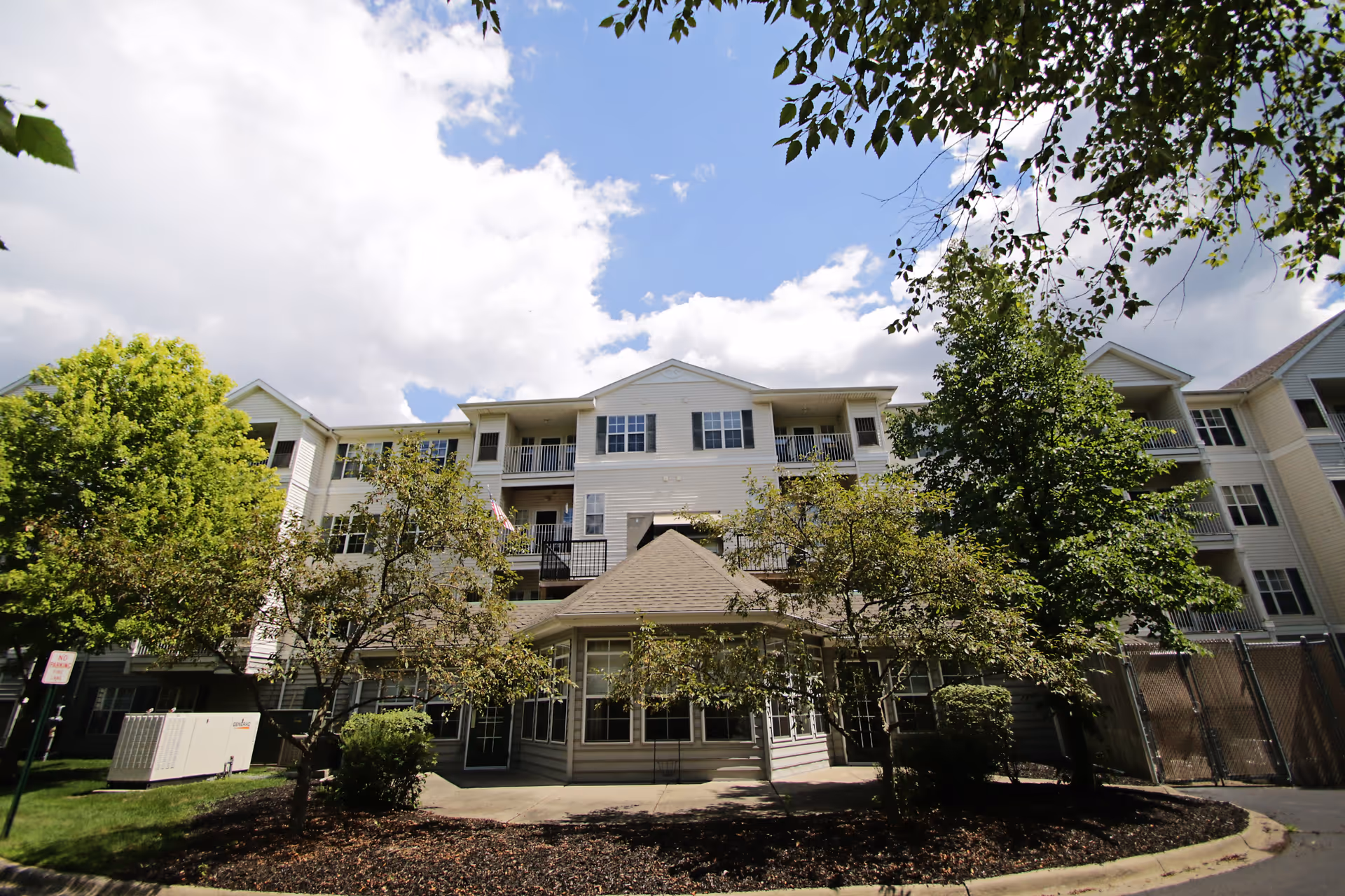 Front exterior of a multi-story senior living building with balconies, trees, and a covered entrance under a partly cloudy sky.