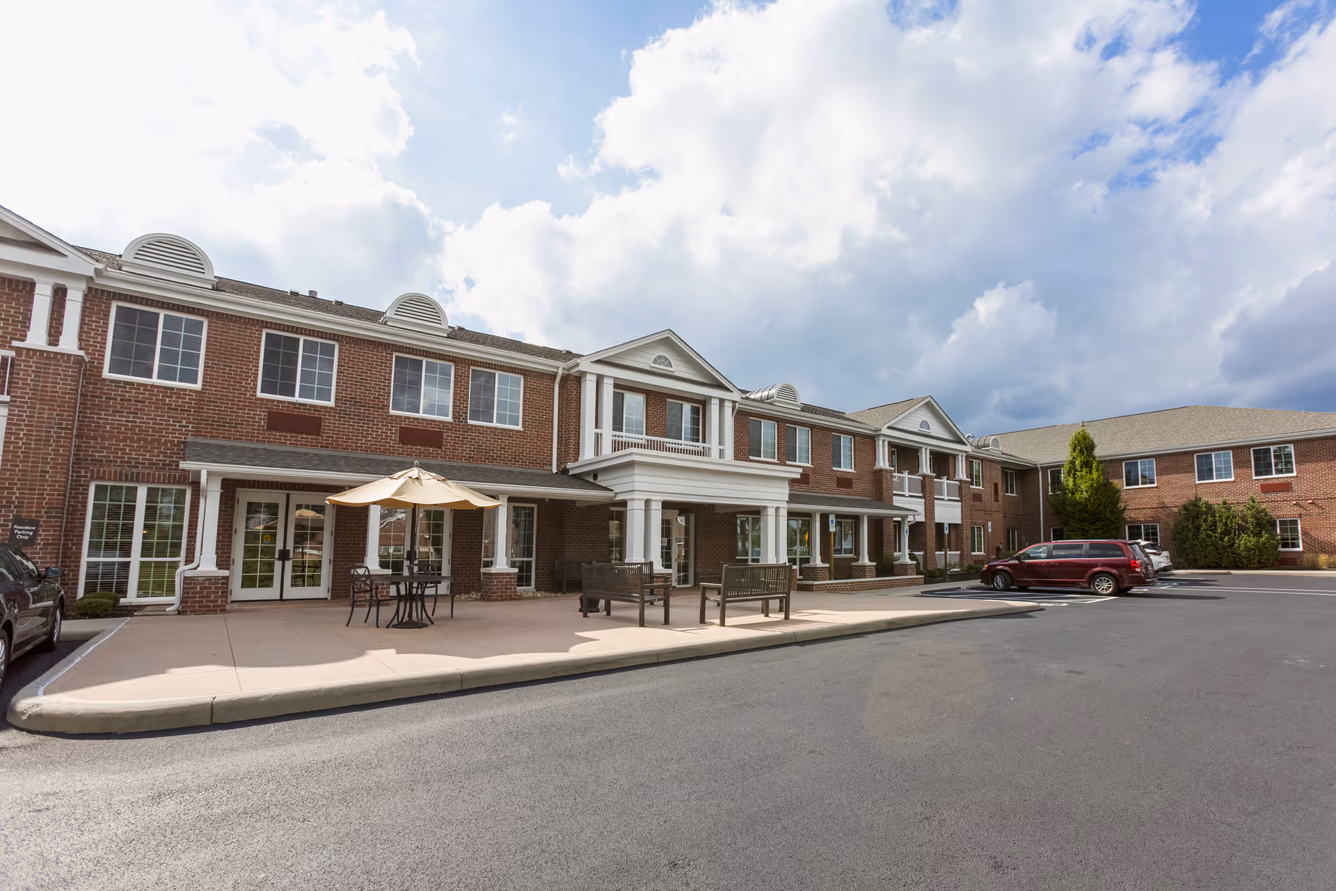 Exterior view of a two-story brick building with multiple windows and a covered entrance area. There are outdoor tables with umbrellas and chairs on the sidewalk in front of the building. Several cars are parked in the parking lot adjacent to the building under a partly cloudy sky.