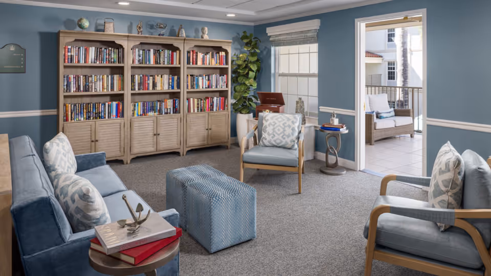 A cozy senior living room with blue walls and carpeted floor featuring a blue sofa with patterned cushions, two matching armchairs, a blue ottoman, and a wooden bookshelf filled with books. A small round side table holds a few books and a decorative item. There is a large window and an open door leading to a balcony with outdoor seating.