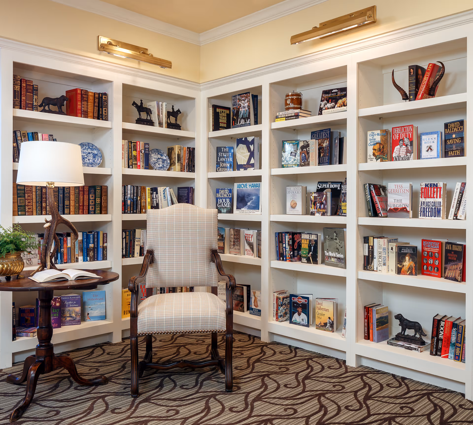 A cozy reading nook featuring a cushioned armchair with wooden arms placed in front of built-in white bookshelves filled with books and decorative items. A round wooden side table with a lamp and an open book sits next to the chair. The room has patterned carpet flooring and warm lighting.