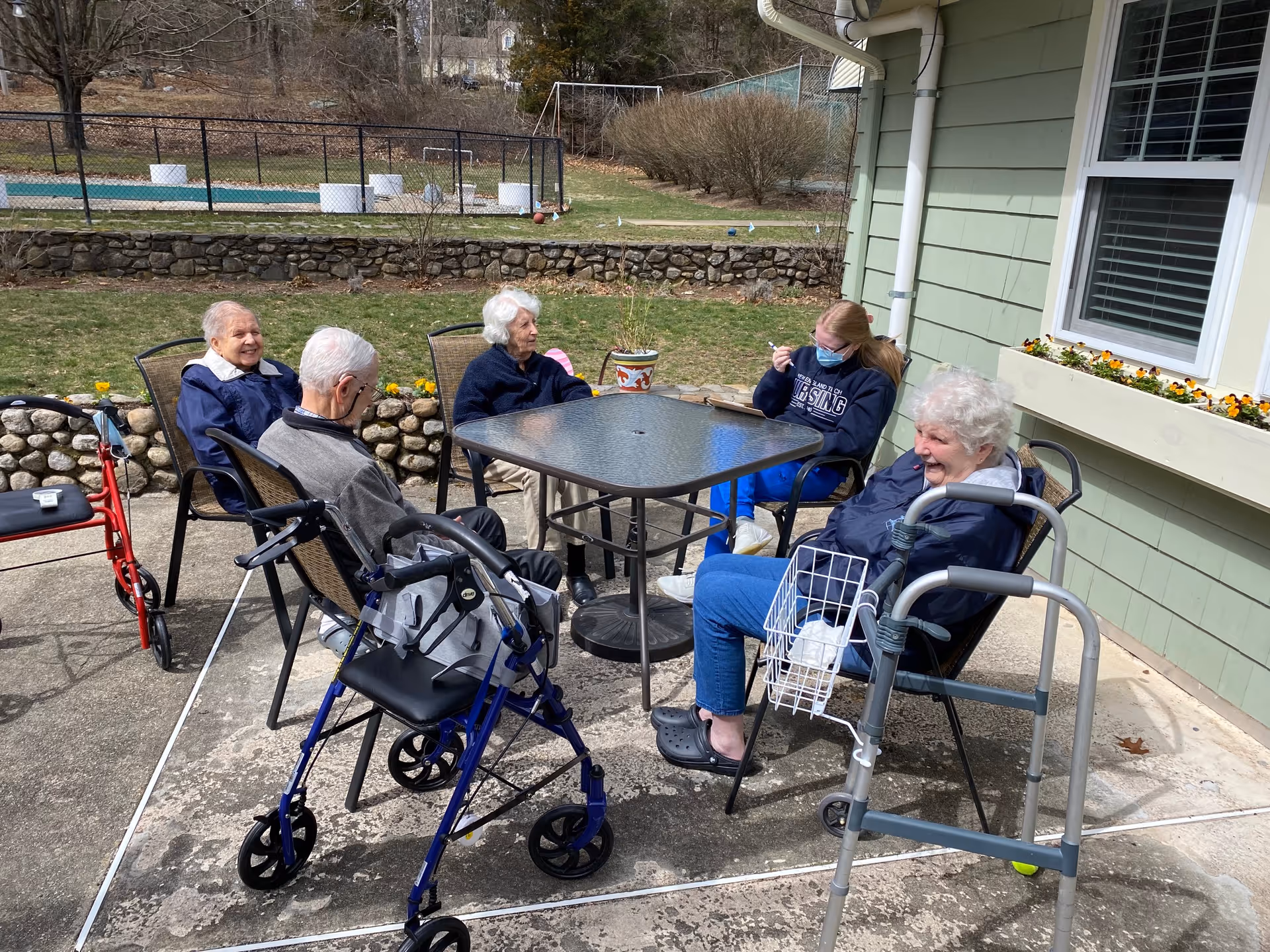 Five elderly individuals and one caregiver sitting around a glass outdoor table on a concrete patio. Two walkers are visible near the group. The setting is outside a green building with a window box of flowers, and there is a stone wall and grassy area in the background.