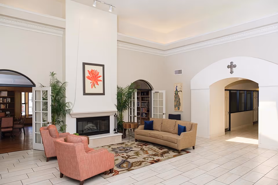 A bright and spacious living room area with a beige sofa and two coral armchairs arranged around a patterned rug in front of a white fireplace. The room features high ceilings with crown molding, two large potted plants flanking the fireplace, and artwork on the walls including a framed red flower above the fireplace and a peacock painting. French doors lead to a room with bookshelves and a table with chairs. There is an arched doorway with a decorative cross above it leading to a hallway.