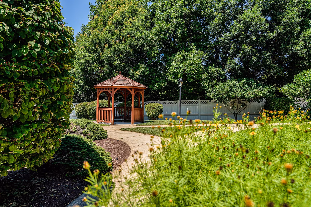 A peaceful outdoor garden area with a wooden gazebo surrounded by lush green trees, bushes, and flowering plants. A paved walkway curves through the garden, and a white fence is visible in the background under a clear blue sky.