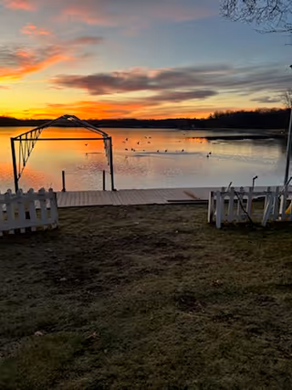 A serene lakeside view at sunset with a wooden dock extending into the water. There is a metal frame structure on the dock and a white picket fence on the grassy shore. Ducks are swimming on the lake, and the sky is filled with colorful clouds reflecting the setting sun.