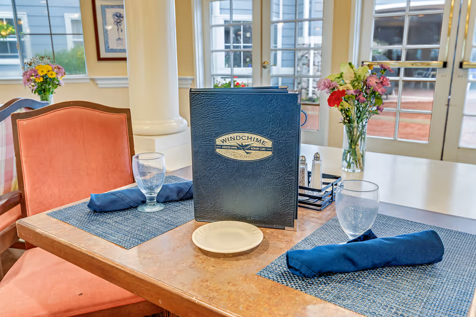 Dining table set for two with a 'Windchime' menu, napkins, glasses, and a vase of flowers in front of glass doors.