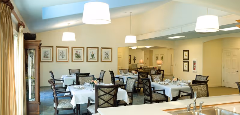 A bright dining room with multiple tables covered in white tablecloths, set with plates, glasses, and silverware. The room features cushioned chairs with patterned upholstery, framed botanical prints on the wall, and large pendant lights hanging from the ceiling. A glass-front cabinet is visible in the corner, and a kitchen counter with a sink is in the foreground.