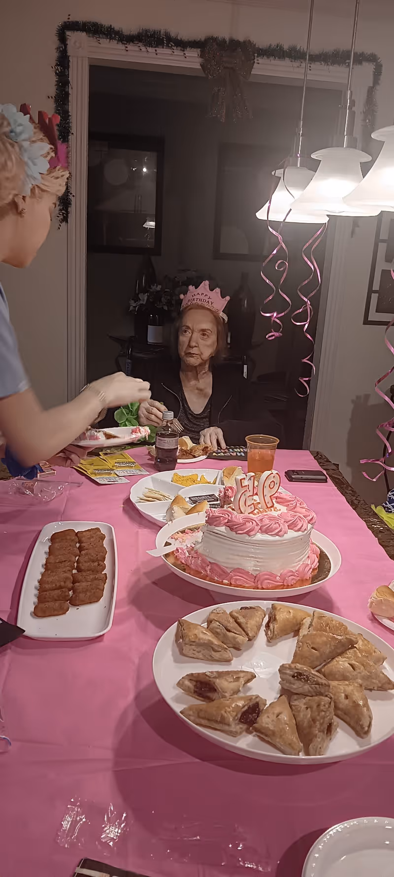 An elderly woman wearing a pink birthday crown sits at a dining table decorated with a pink tablecloth, a pink and white birthday cake with candles, plates of snacks, and drinks. A person on the left side of the image is serving or handing something to the woman. Pink spiral streamers hang from the ceiling lights above the table.