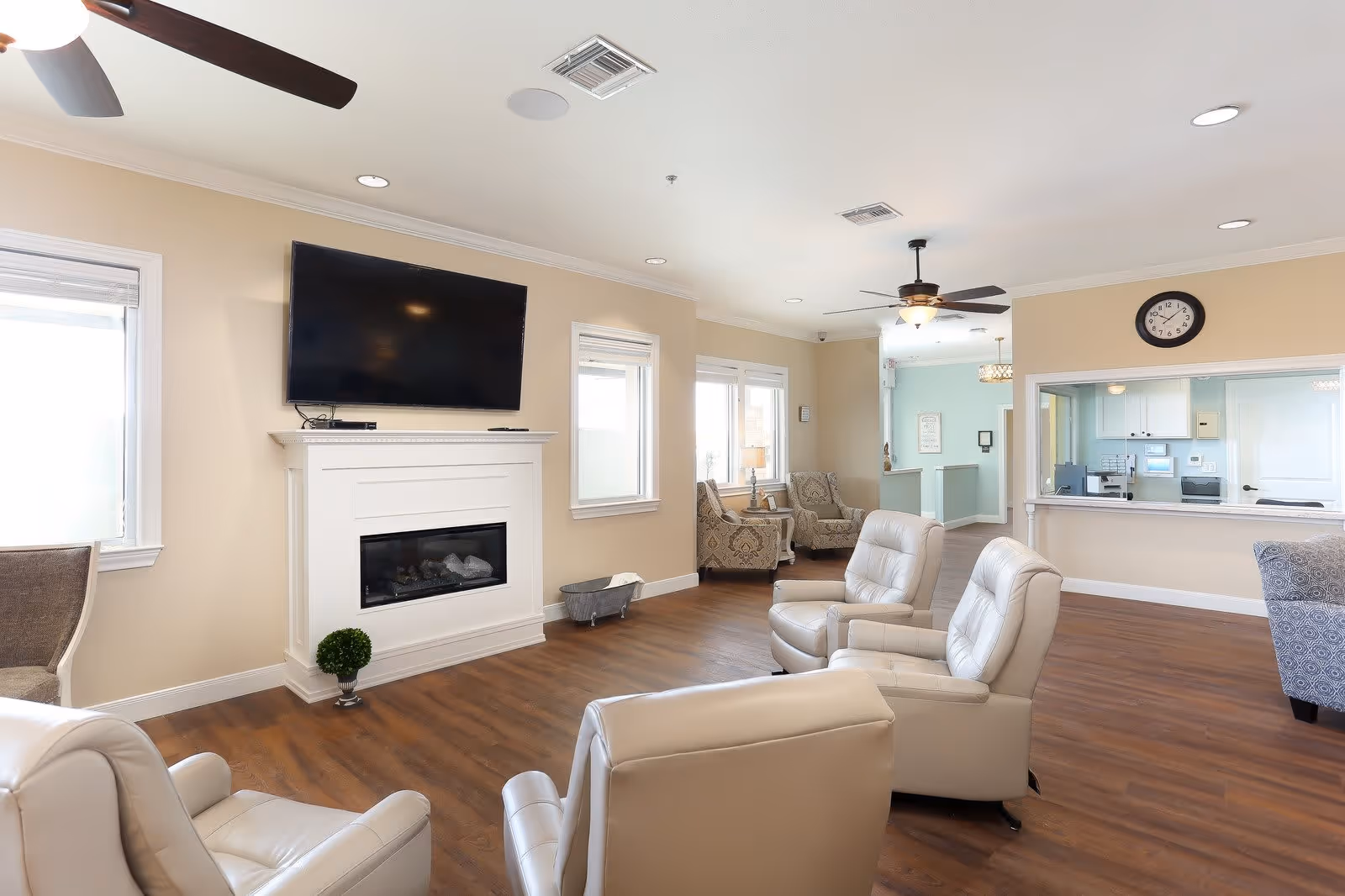 Bright common room with leather recliners facing a wall-mounted TV above a fireplace, additional seating, and a reception/service window.