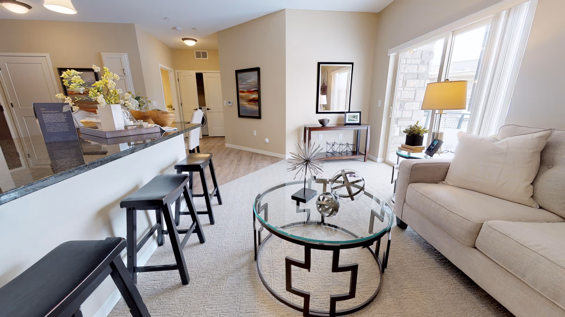 A bright and modern living room area with a beige sofa, a glass coffee table with decorative metal spheres, and a side table with a lamp and plant. There are three black bar stools along a kitchen counter with a vase of flowers and a tray. The room has light-colored walls and carpet, with large windows letting in natural light.