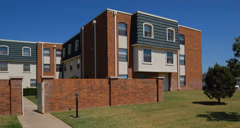 Exterior view of a multi-story senior living facility building with brick and light-colored walls, greenish-blue siding on the upper floors, and several windows. The building is surrounded by a well-maintained lawn, a brick wall, a sidewalk, and a few trees under a clear blue sky.