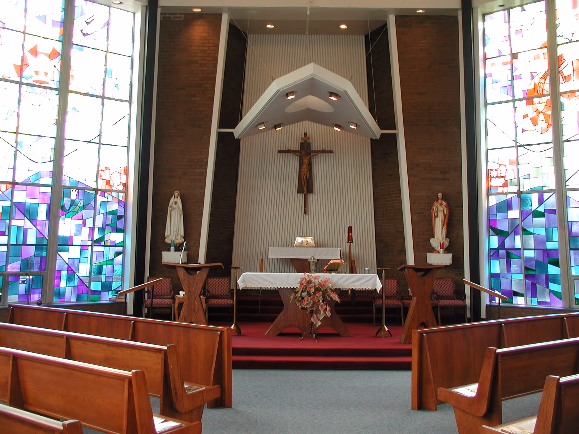 Interior view of a chapel with wooden pews, stained glass windows on both sides, a crucifix on the wall behind the altar, and statues on either side of the altar.
