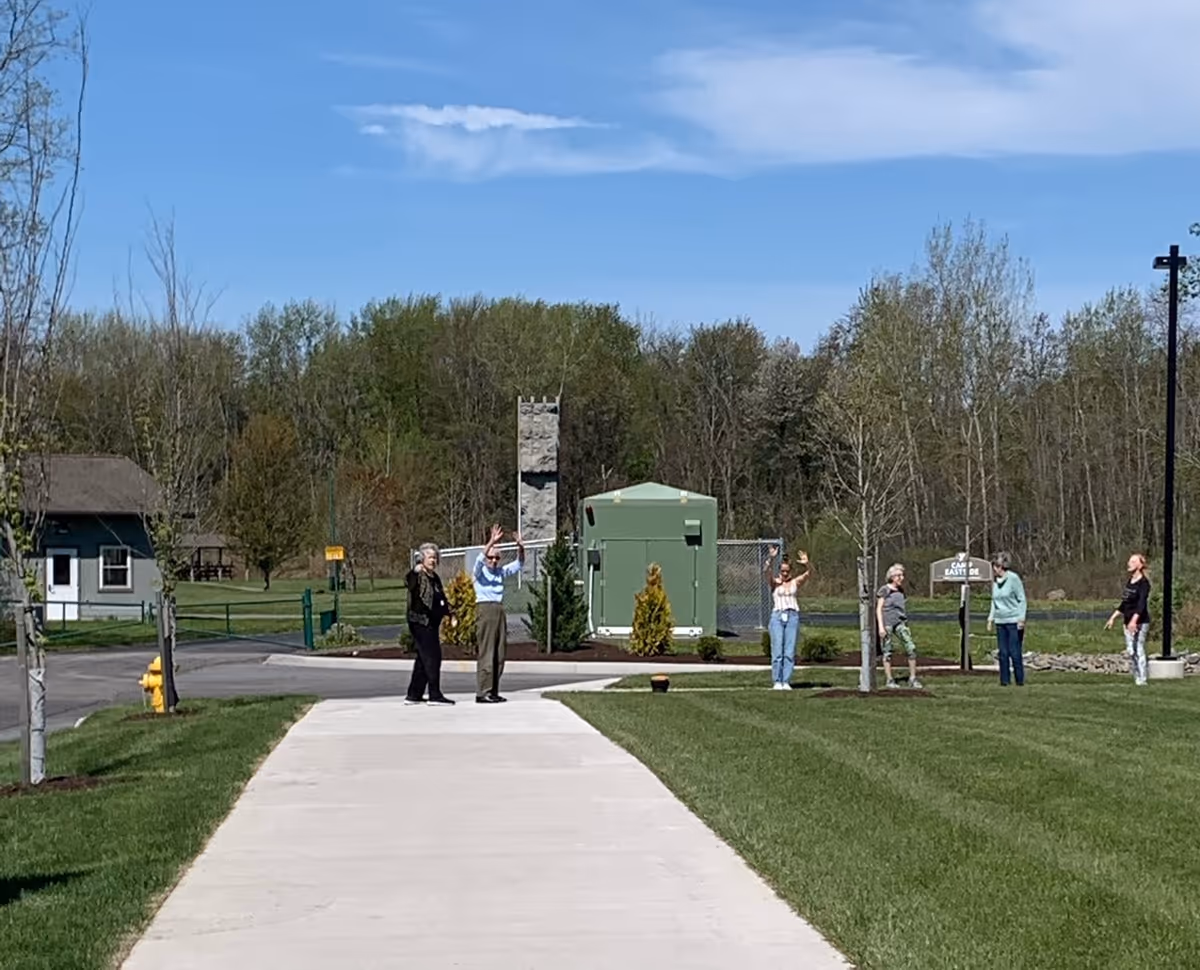 Several people standing and waving on a paved walkway outside a senior living community, with lawns, trees, and utility buildings in the background.