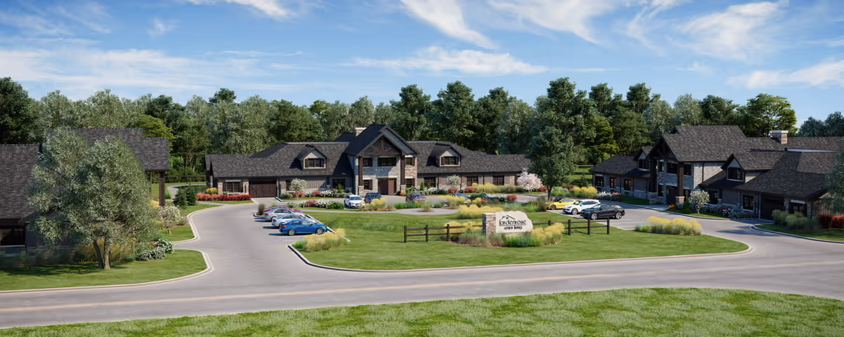 Exterior view of a landscaped senior living campus with multiple lodge-style buildings, a circular driveway, parked cars and a central entrance sign.