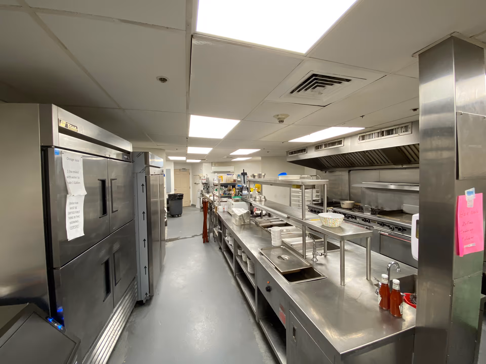 A commercial kitchen with stainless steel appliances and countertops, including large refrigerators on the left and cooking ranges with exhaust hoods on the right. The kitchen is well-lit with ceiling lights and has various kitchen utensils and containers placed on the counters.