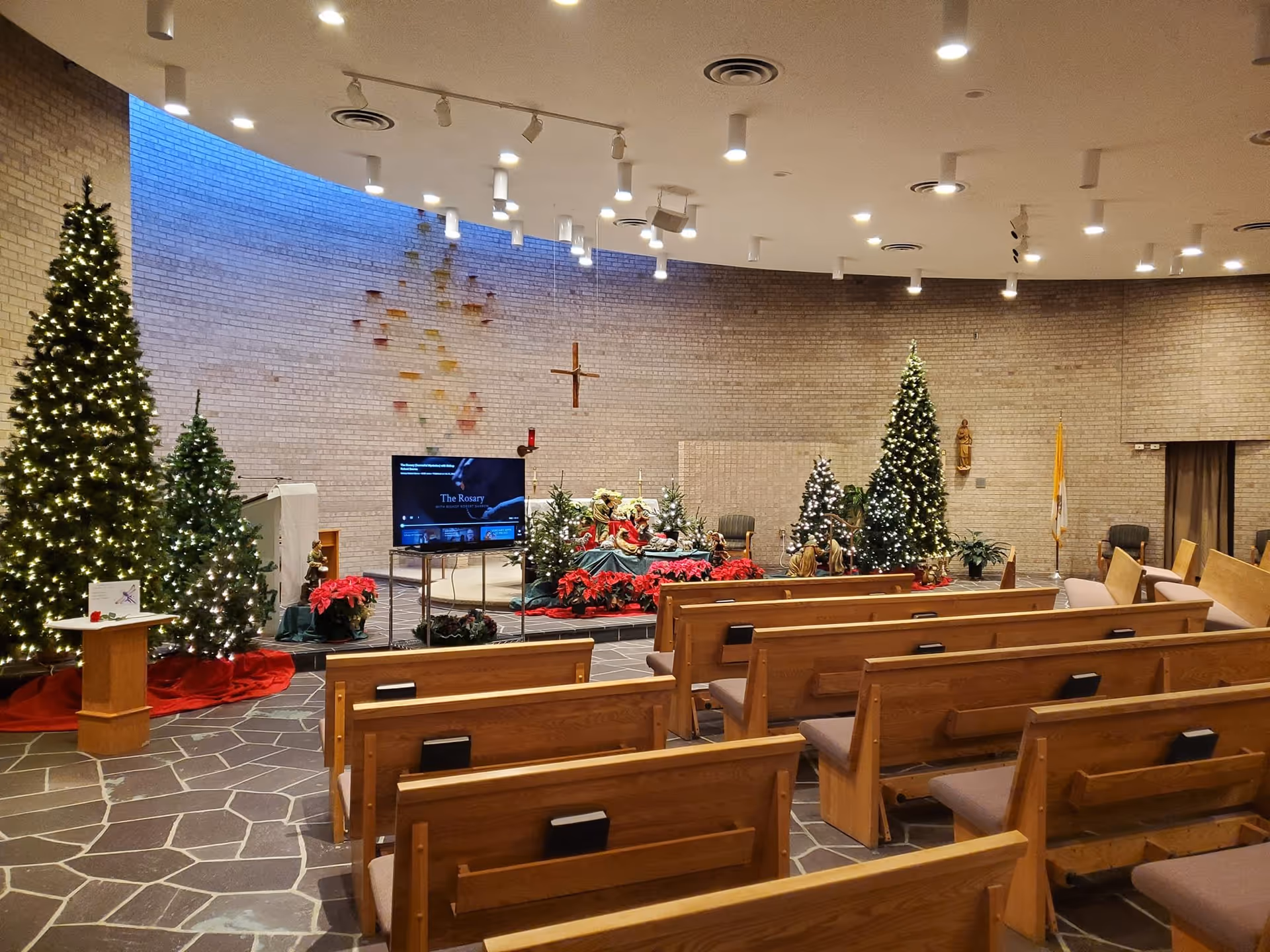 Interior of a chapel decorated for Christmas with several lit Christmas trees, poinsettias, a nativity scene, wooden pews, a cross on a brick wall, and a TV screen displaying 'The Rosary'.