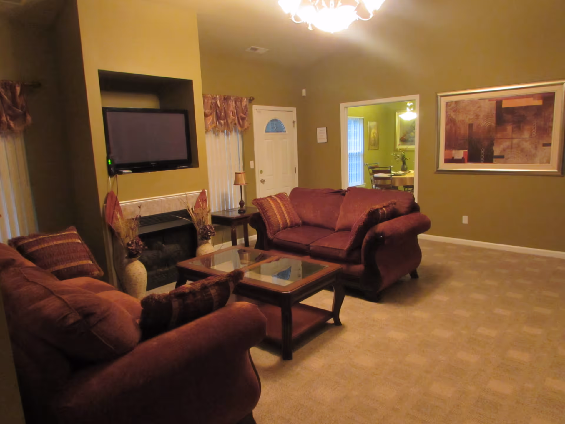 A cozy living room with two maroon upholstered sofas facing each other, a glass-top coffee table in between, a wall-mounted flat-screen TV above a fireplace, and a large abstract painting on the wall. The room has beige carpet, greenish walls, and a doorway leading to a dining area with a table and chairs.