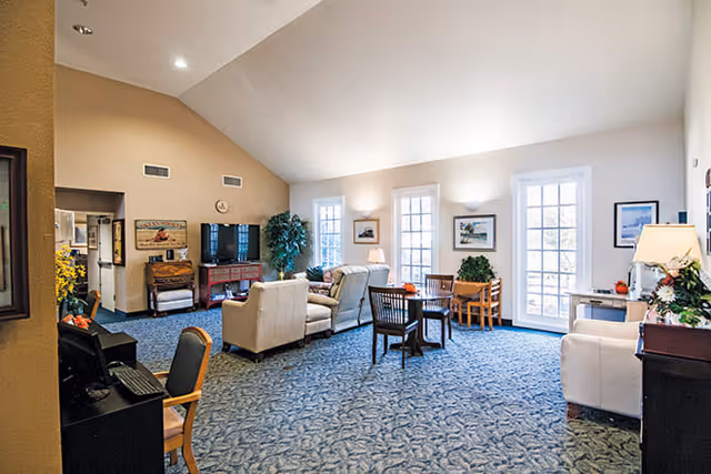 A spacious senior living common area with blue patterned carpet, beige walls, and high ceilings. The room features several seating arrangements including beige sofas and wooden chairs around small tables. There is a television on a wooden stand against one wall, framed artwork on the walls, and large windows and glass doors letting in natural light. Potted plants and lamps add to the cozy atmosphere.