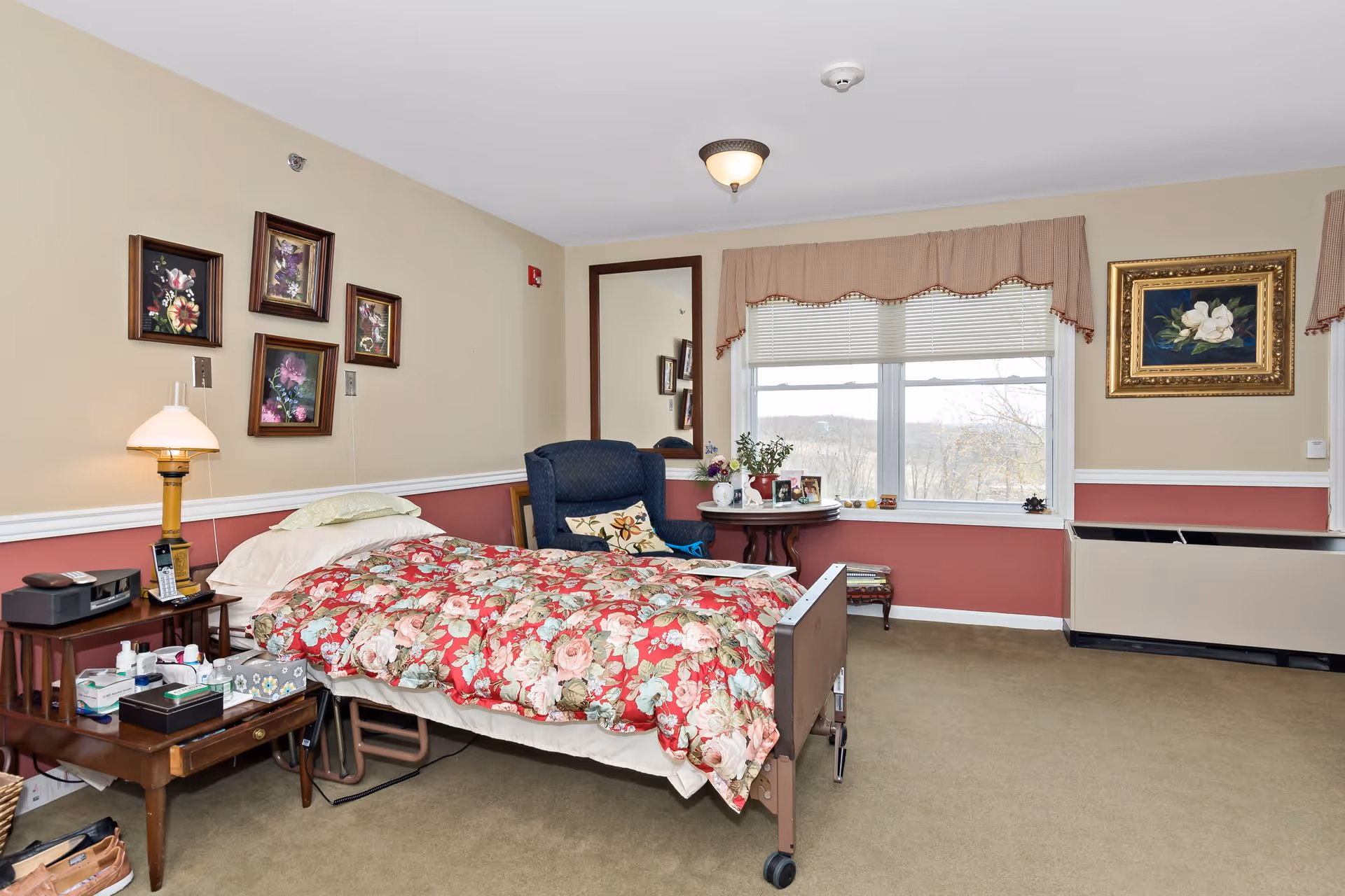 A cozy assisted living bedroom with a single bed covered in a red floral comforter. Next to the bed is a wooden nightstand with a lamp, phone, and various personal items. A blue armchair with a floral pillow sits beside a round table holding framed photos and a plant. The room has beige and red walls adorned with framed floral paintings, a large mirror, and a window with blinds and a valance overlooking an outdoor view.