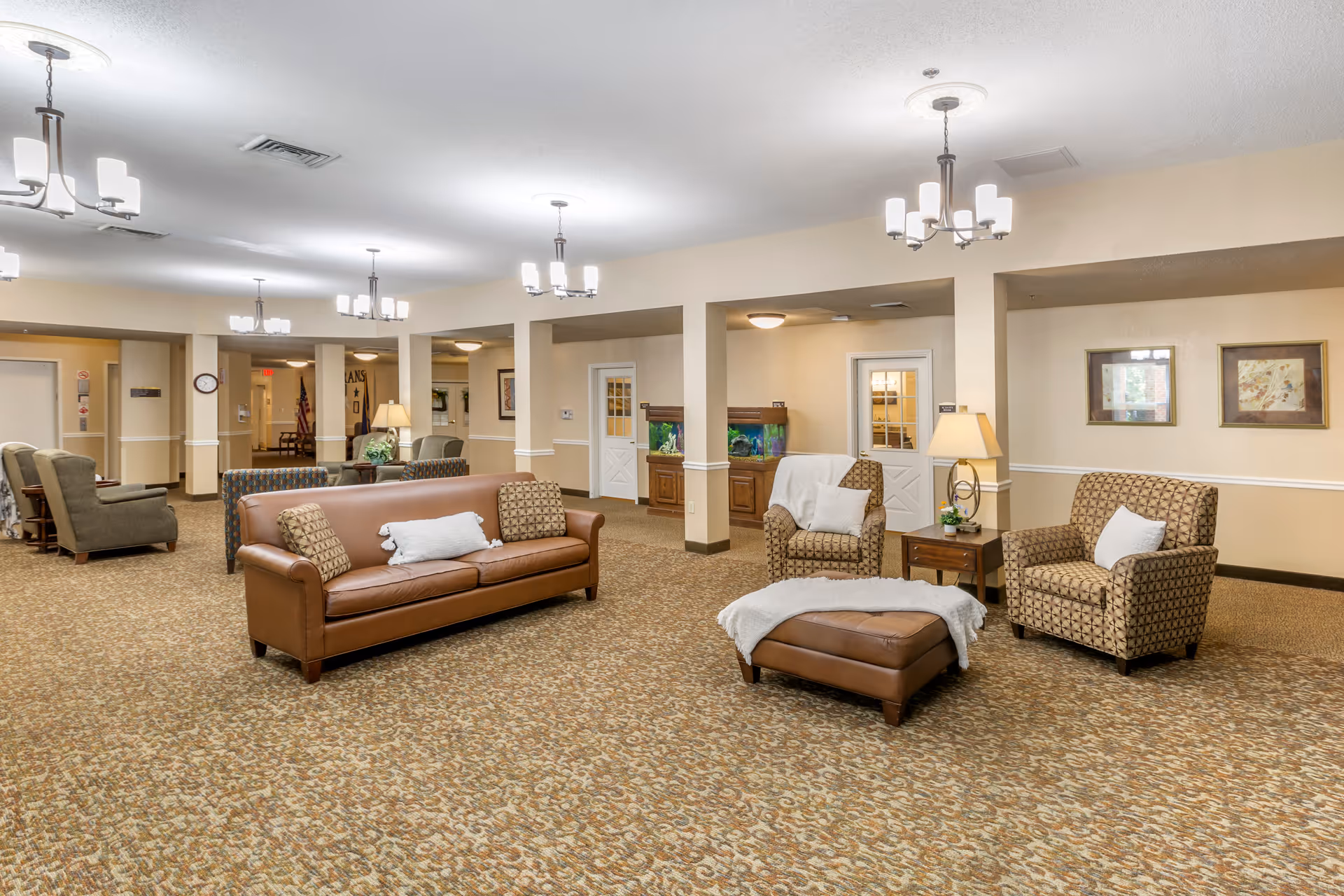 A spacious senior living facility common area with beige walls and patterned carpet. The room features a brown leather sofa with pillows, two patterned armchairs with white pillows, a matching ottoman with a white throw, a wooden side table with a lamp, and an aquarium in the background. Several ceiling light fixtures illuminate the space, and framed artwork hangs on the walls.
