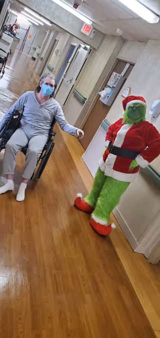A person wearing a face mask sits in a wheelchair in a hallway of a care facility. Next to them stands someone dressed in a Grinch costume wearing a Santa Claus outfit. The hallway has wooden flooring, beige walls, handrails, and several doors along the corridor.