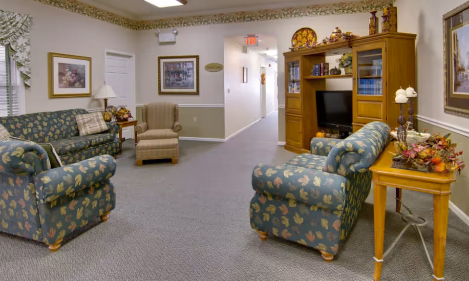 Carpeted senior living common room with patterned sofas and armchairs, a wooden entertainment center, and decorative accents.
