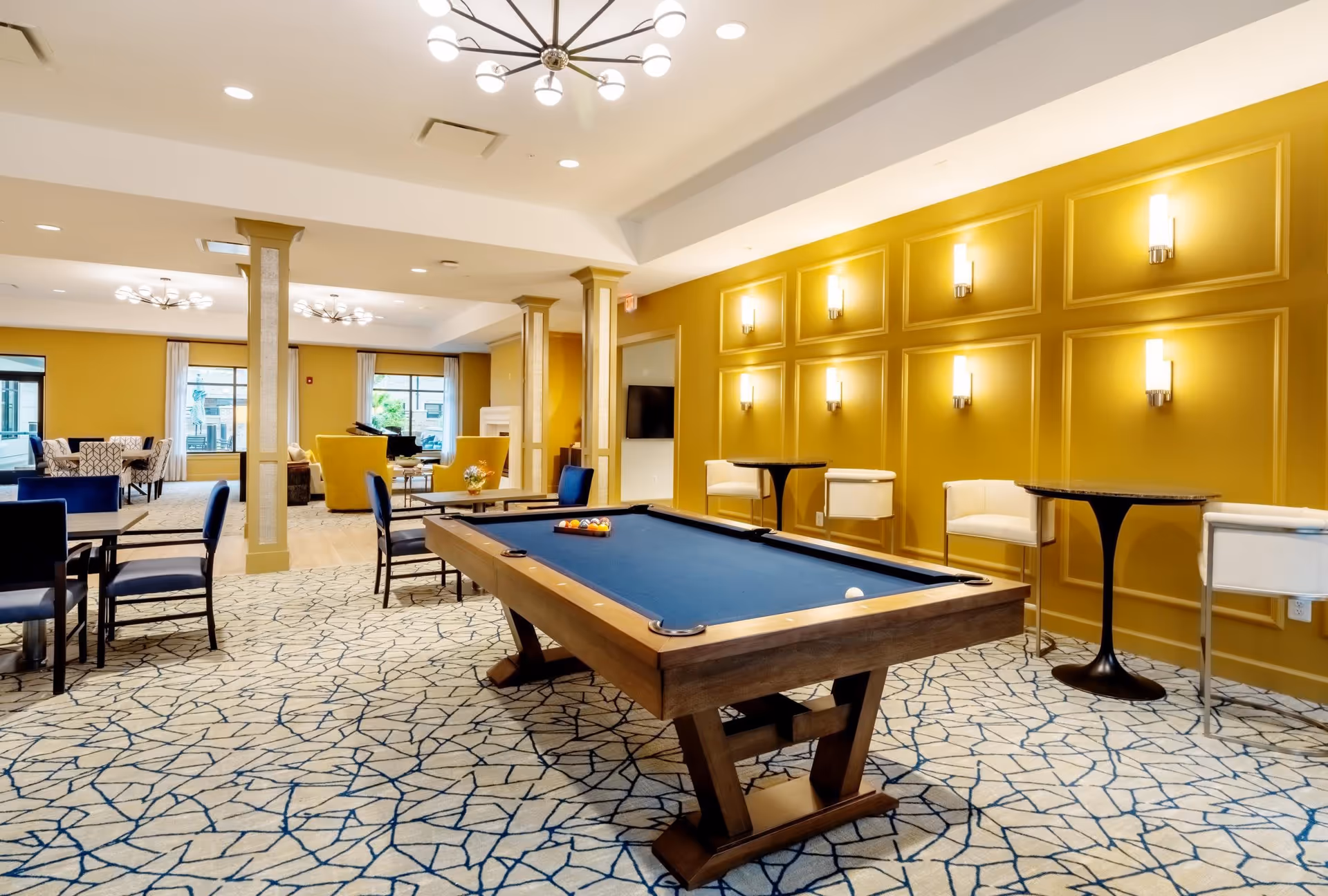 A spacious and well-lit common area featuring a pool table with blue felt in the center, surrounded by chairs and small tables. The room has mustard yellow walls with decorative paneling and modern wall sconces. In the background, there are additional seating areas with armchairs and large windows letting in natural light.
