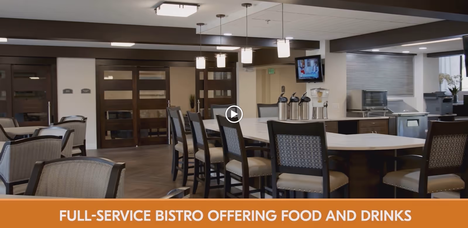Interior view of a full-service bistro area in a retirement community featuring a large central island with chairs, coffee dispensers, and modern lighting fixtures. There are additional seating areas with tables and chairs, and a television mounted on the wall.
