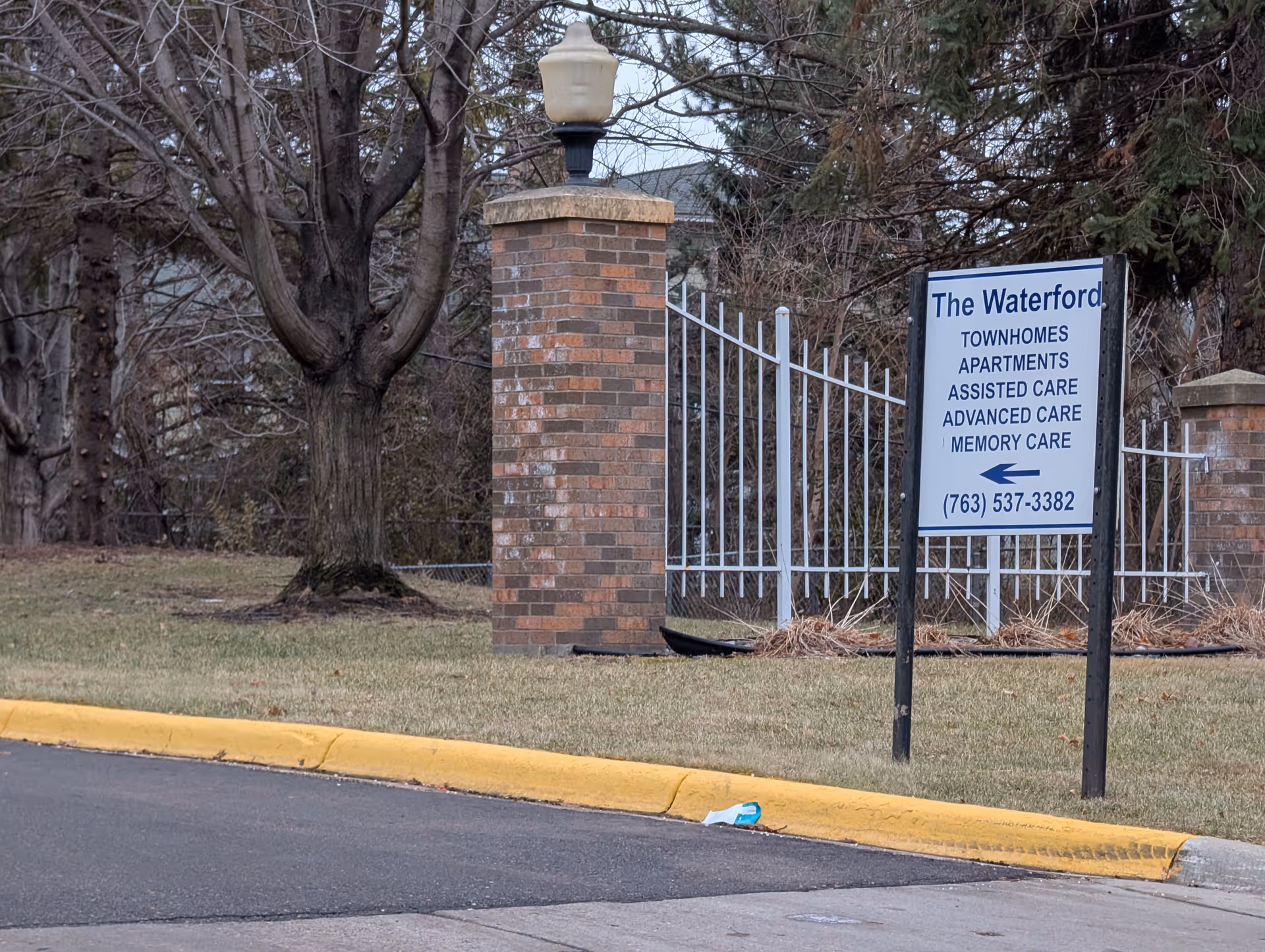 Outdoor view of a sign for The Waterford facility, listing townhomes, apartments, assisted care, advanced care, and memory care services. The sign is placed on a grassy area near a brick pillar and white metal fence, with leafless trees in the background.