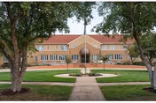 Front exterior view of Newton Presbyterian Manor, a two-story brick building with a red roof, large windows, and a central entrance. The building is framed by two large trees and a circular driveway with a fountain in the center.
