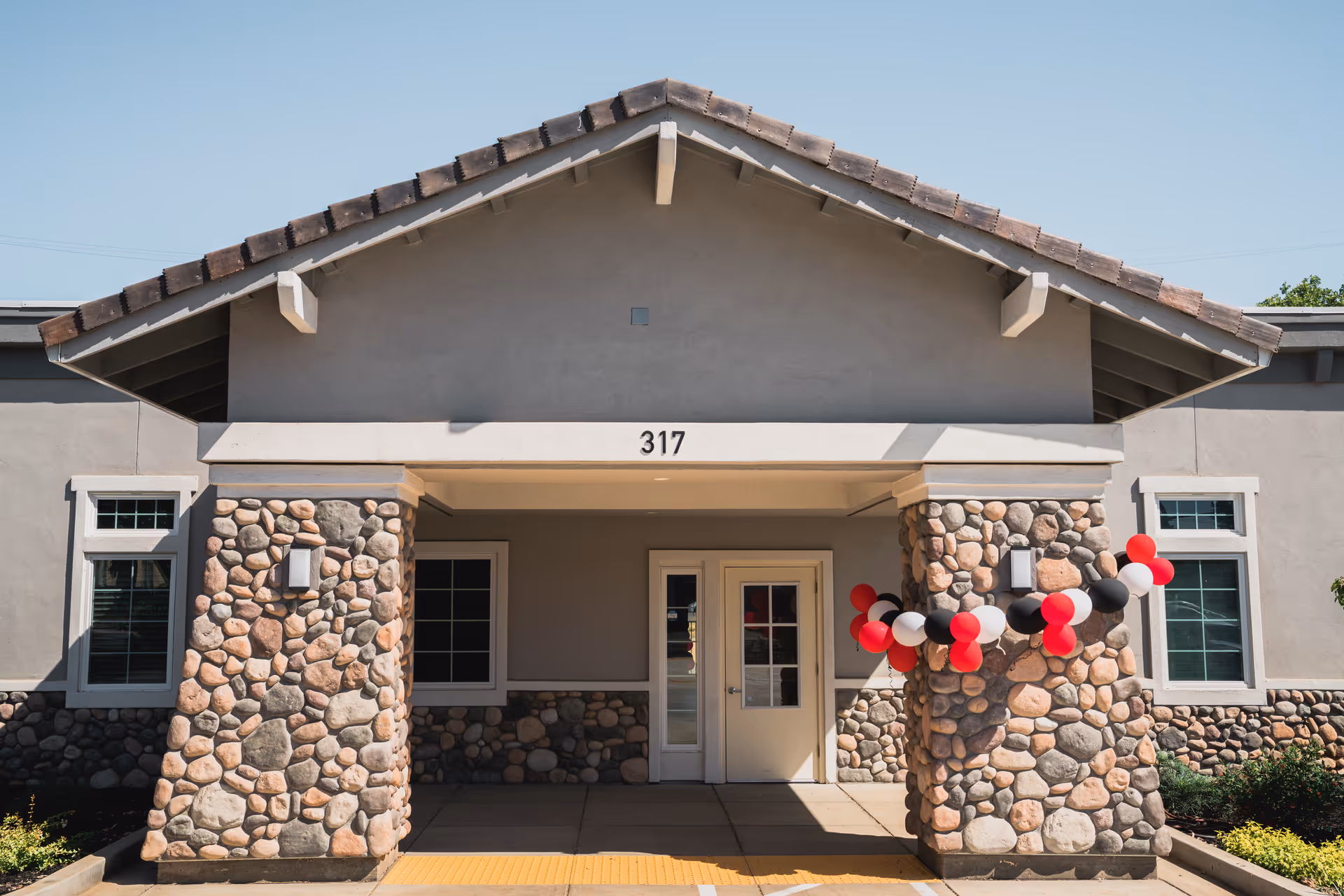 Front exterior view of a building with stone pillars and a gabled roof. The building has the number 317 above the entrance. Red, black, and white balloons are tied to the right stone pillar.