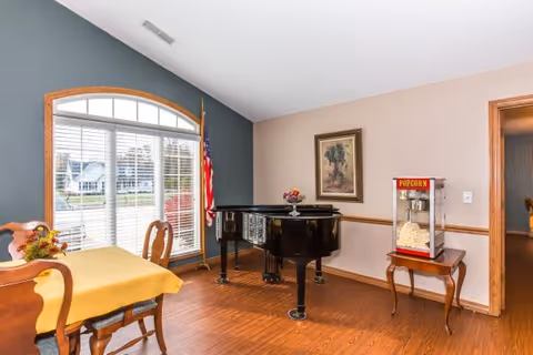 A bright room with a large window featuring white blinds and an American flag beside it. There is a wooden table with a yellow tablecloth and chairs on the left side. In the center of the room is a black grand piano with a decorative bowl on top. On the right side, a small wooden table holds a popcorn machine. The walls are painted in two tones, with one side gray and the other beige, and a framed picture hangs above the popcorn machine.