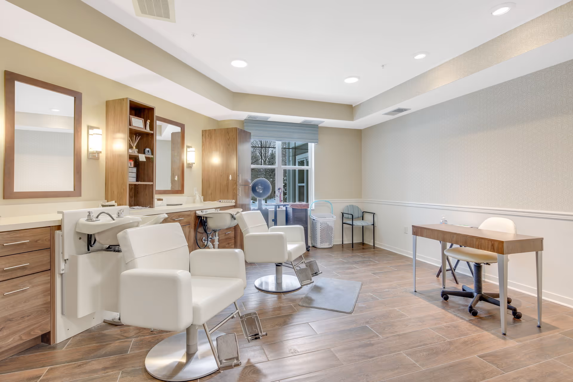 A bright and clean salon area in a senior living facility featuring two white salon chairs with attached sinks, wooden cabinetry with mirrors and lights above, a small desk with a chair on the right side, and a window letting in natural light in the background.