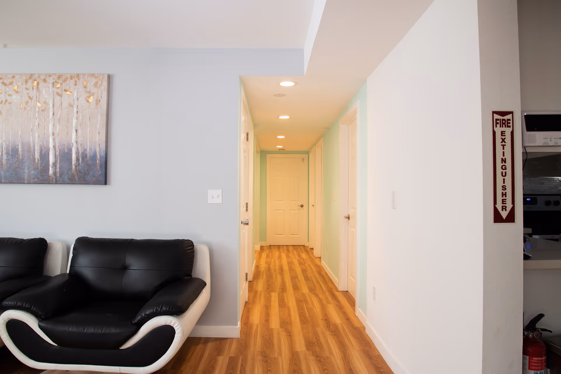 Interior view of a hallway in a senior living facility with wooden flooring and white doors. To the left, there is a black and white cushioned chair and a wall painting depicting abstract trees. On the right wall, there is a fire extinguisher sign and a glimpse of a kitchen area with appliances.