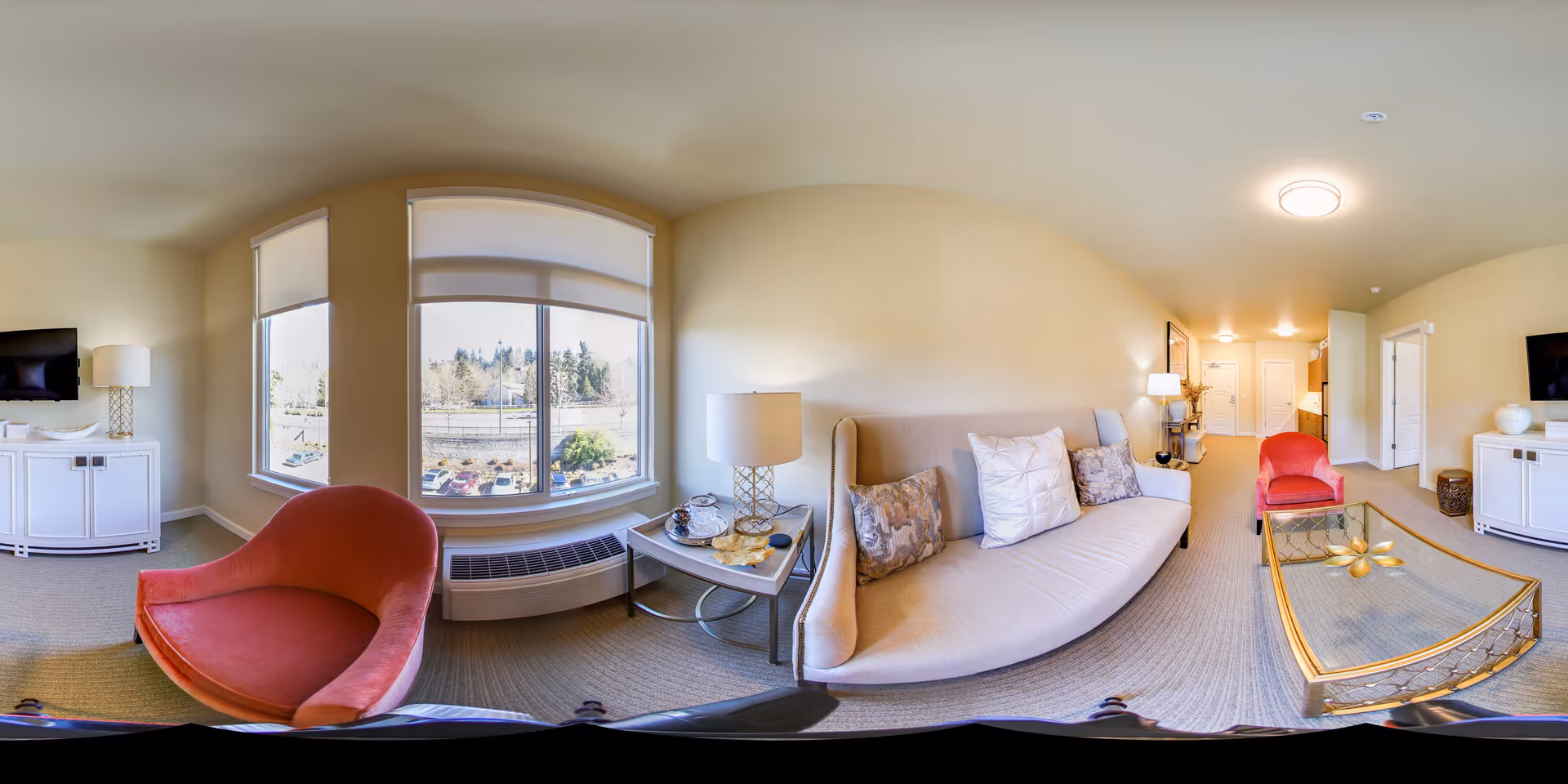 Panoramic living room with a beige sofa, two red accent chairs, a glass coffee table, TV cabinets, and large windows overlooking greenery.