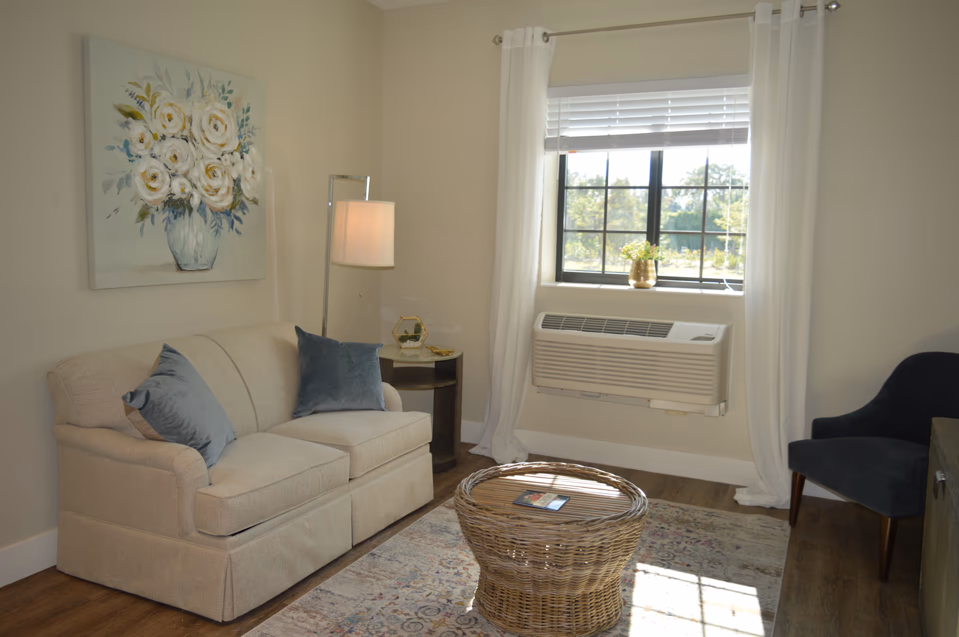 A cozy living room with a beige loveseat adorned with two blue throw pillows, a wicker coffee table on a patterned rug, a dark blue armchair, a side table with a lamp, and a window with white curtains and blinds letting in natural light. A painting of white flowers in a vase hangs on the wall above the loveseat.