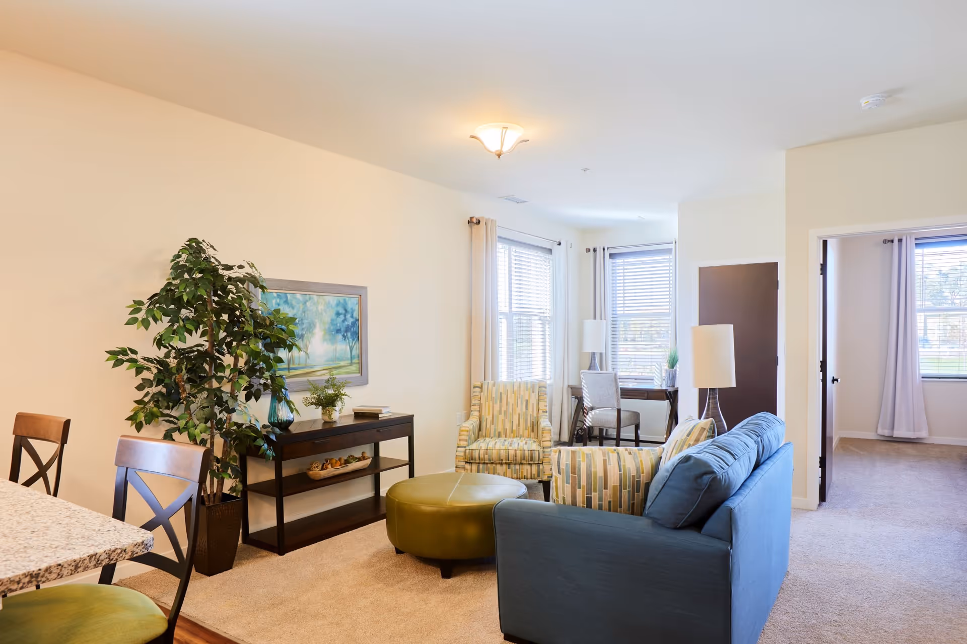 A bright furnished living room with a blue sofa, patterned armchairs, a green ottoman, console table, and dining chairs by sunlit windows.